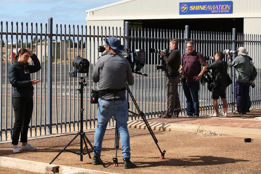A journalist stands before a camera ready to go live next to several photographers looking through a fence at an airport.