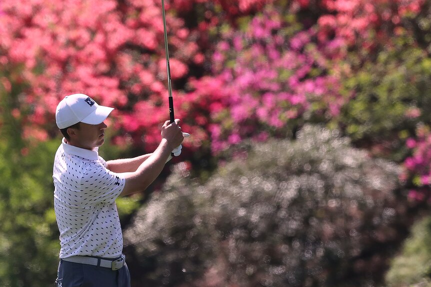 Standing in front of blooming pink azaeleas, Jordan Spieth completes a shot.