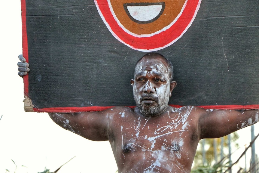 An Indigenous man, his face covered in paint, holds a sign during a dance performance.