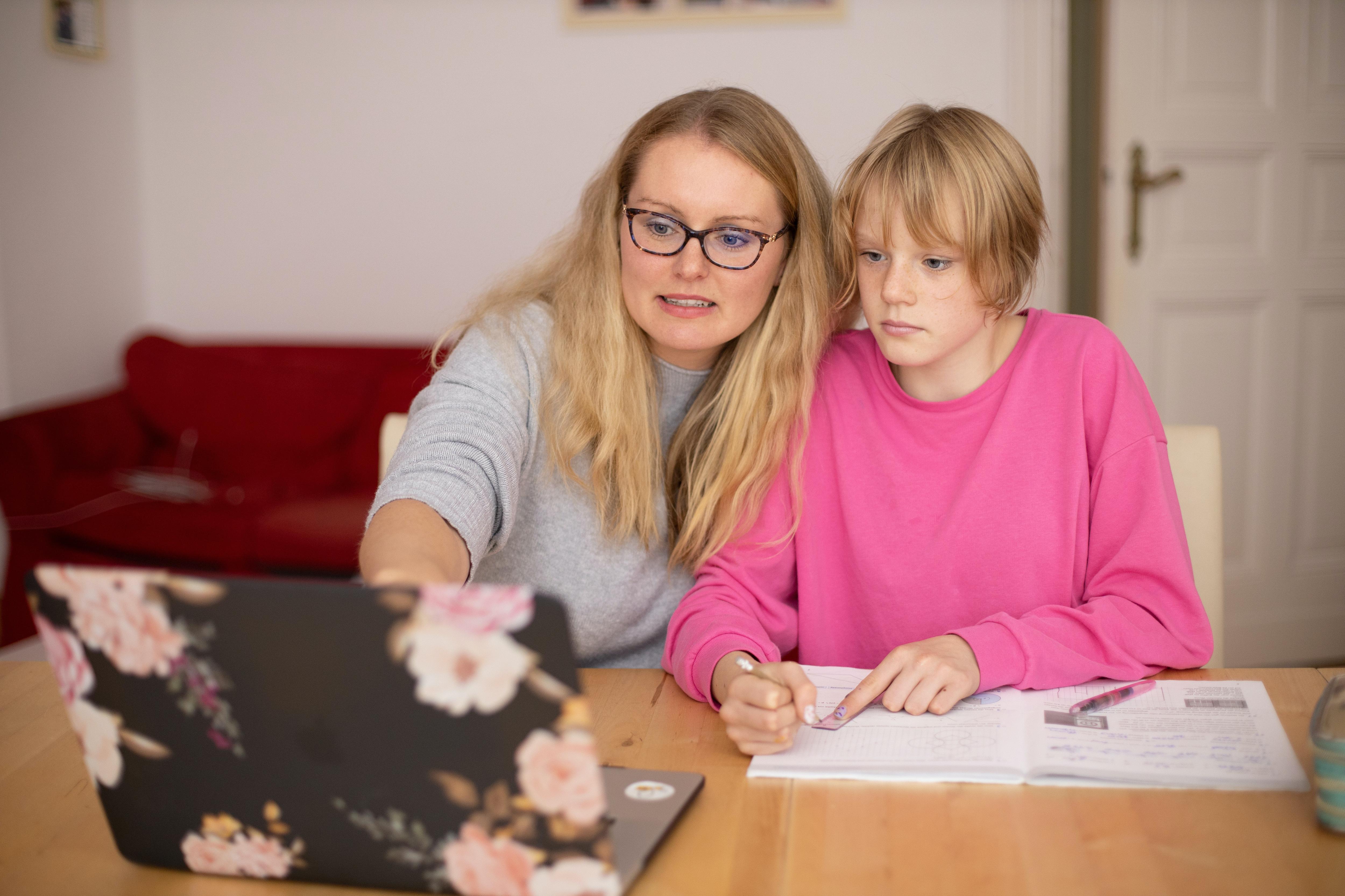A woman with long, fair hair sits close to a teenager with short hair and pink jumper. Both look into a lap-top screen.