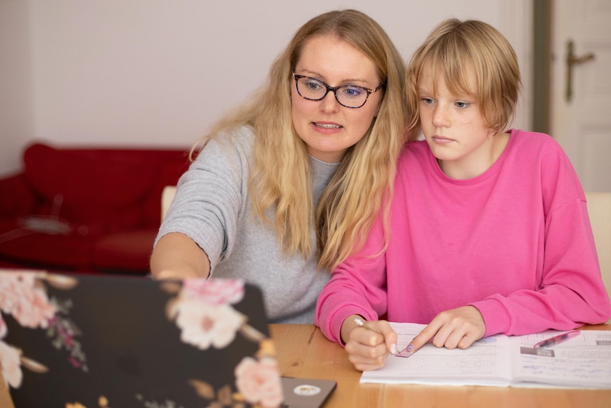 A woman with long, fair hair sits close to a teenager with short hair and pink jumper. Both look into a lap-top screen.