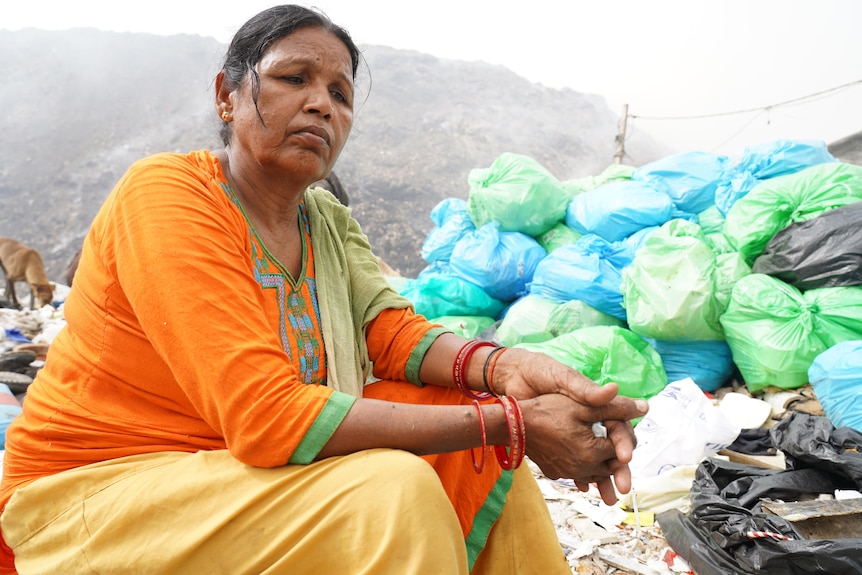 An Indian woman in a vibrant orange sari sits near a huge pile of steaming rubbish, looking exhausted 