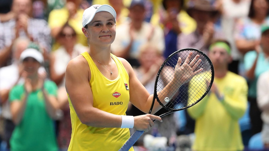 A woman wearing yellow sports singlet and white cap smiles holding tennis racket, while blurred crowds stands clapping behind.