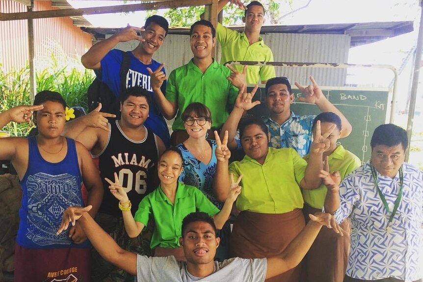 A smiling Australian woman stand among a group of Samoan young people at a school.