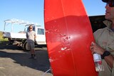 A red surfboard with bite marks from a shark is held up by a man with another man in the background.