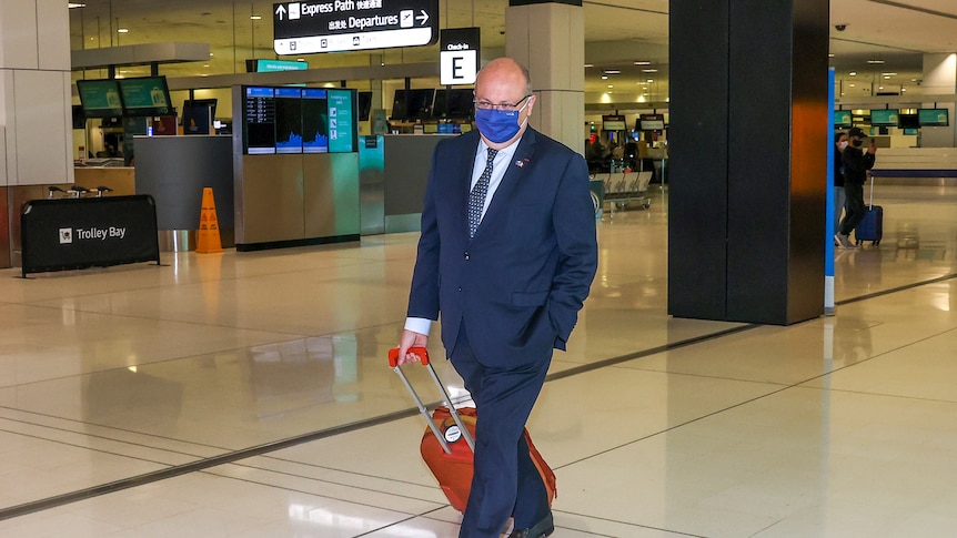 A man wearing a suit and face mask wheels his suitcase in an airport terminal