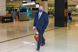 A man wearing a suit and face mask wheels his suitcase in an airport terminal