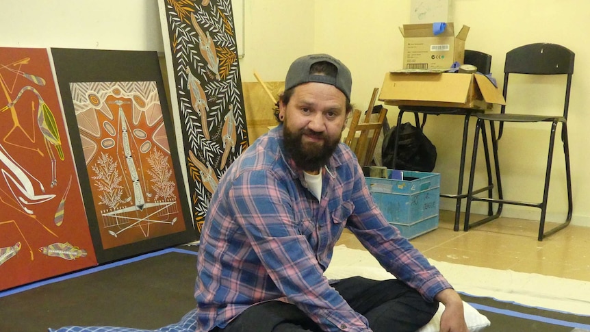 A man wearing his cap backwards and a checked shirt sits in a cushion in front of painting.