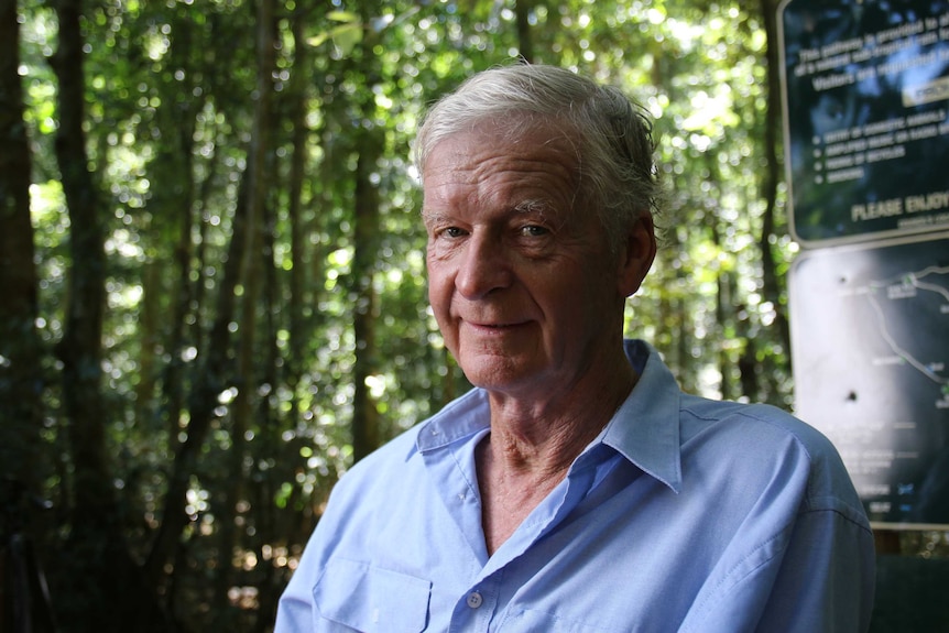 A close up of an older man with grey hair, stands in front of trees, wears blue shirt, looks at the camera.