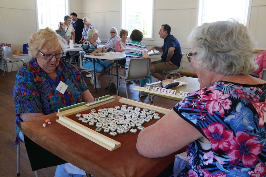 Two senior women with name tags play mah jong in sunlit recreation room with other seniors behind.