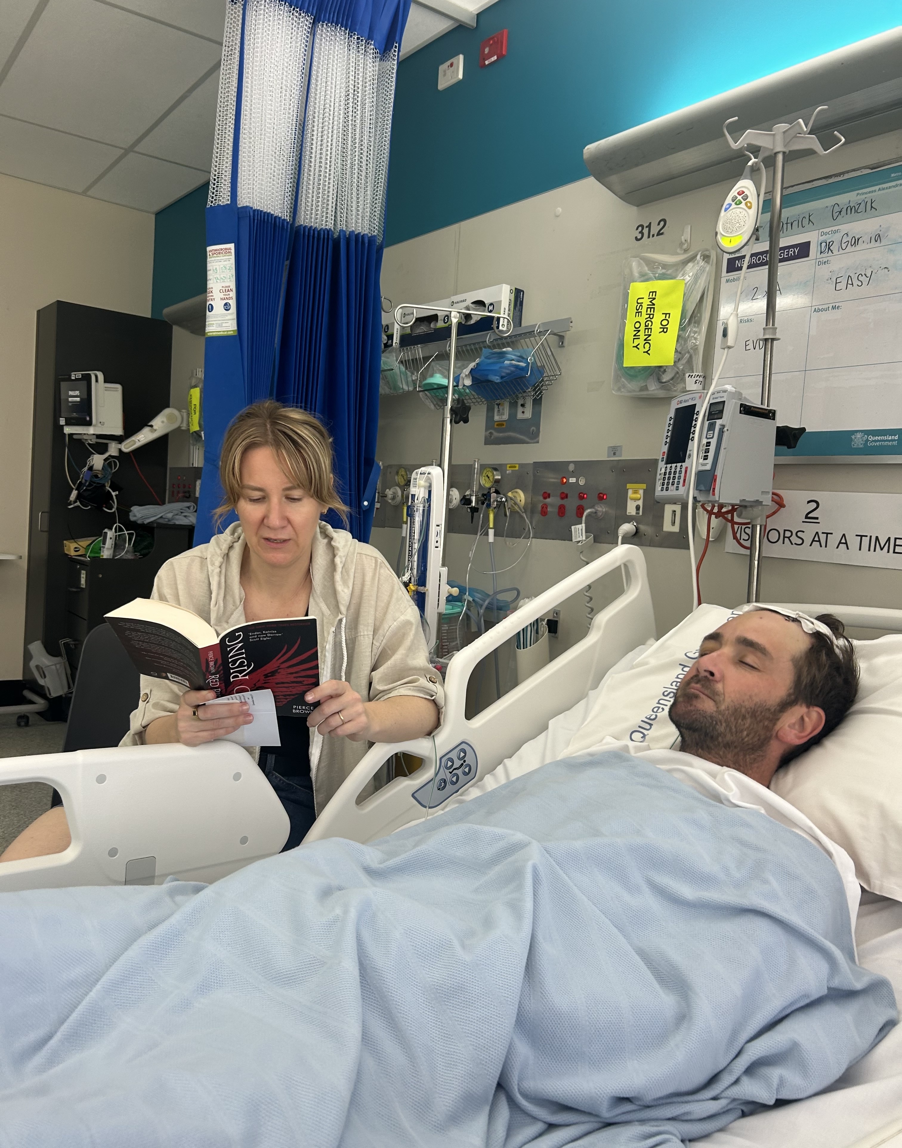 A man lays unconscious in a hospital bed while a woman sits next to him reading.
