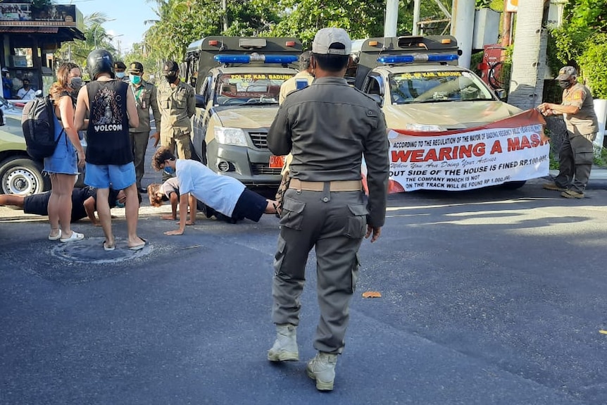 Three men doing push-ups in the street next to local police car with a banner saying wearing a mask.