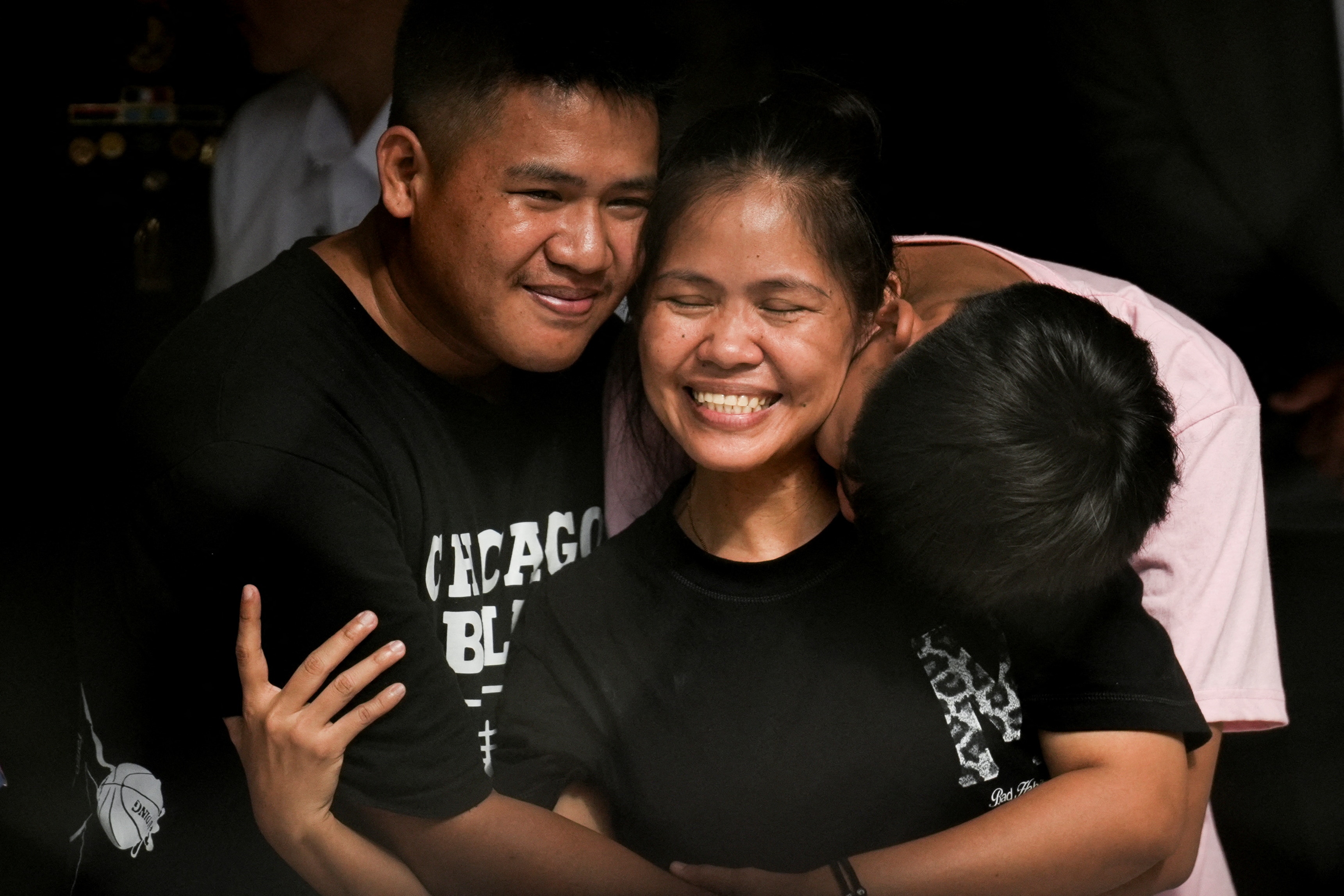A woman smiles as she hugs two young adult men.