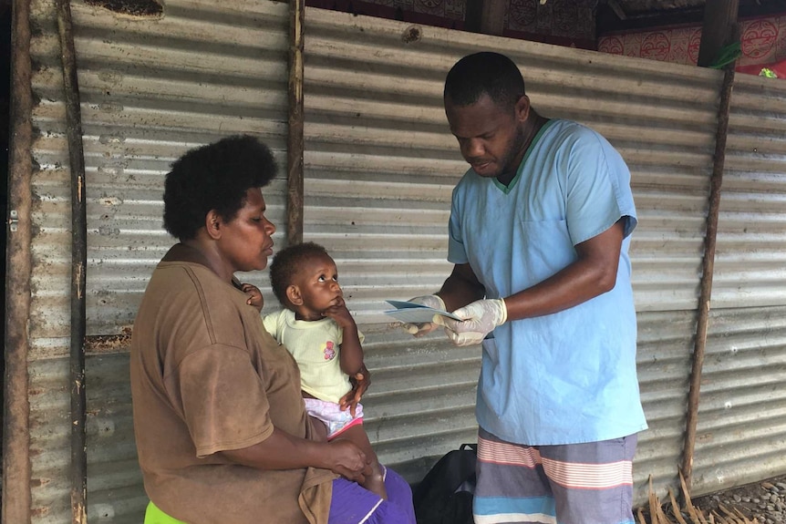 A child looks up at a health worker as she sits on her mother's lap outside a corrugated iron wall in South River, Vanuatu