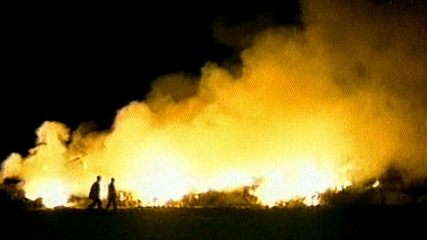 Two people walk at night in front of large glowing controlled blaze