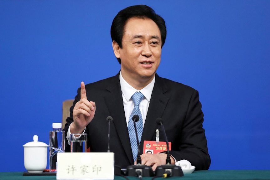 Man in a suit and blue tie during a press conference holding one finger n the air.