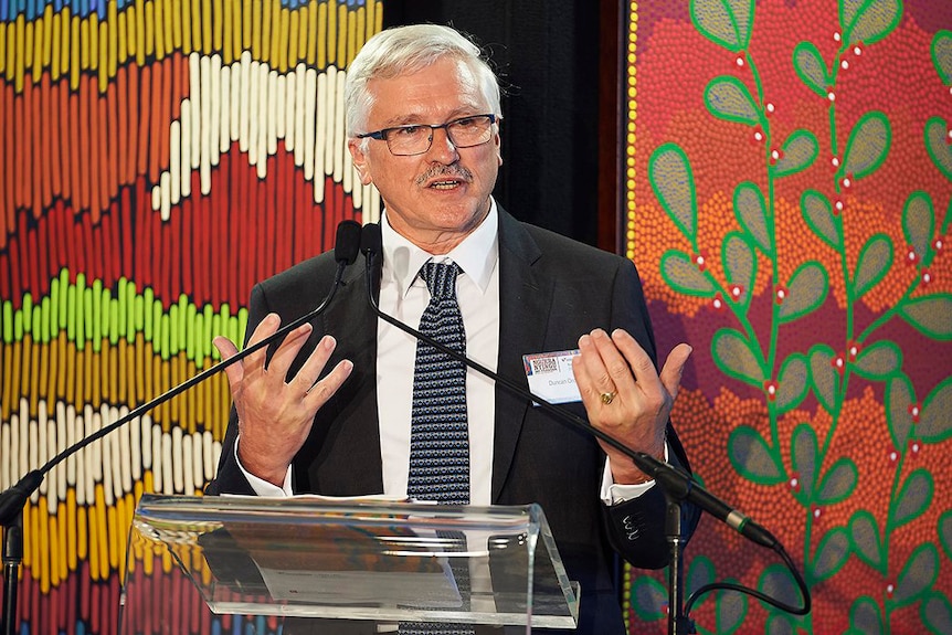 A man stands at a lectern in front of an Indigenous art print.