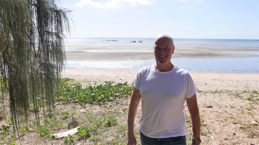A man in a white T-shirt, smiling in front of a tropical beach.