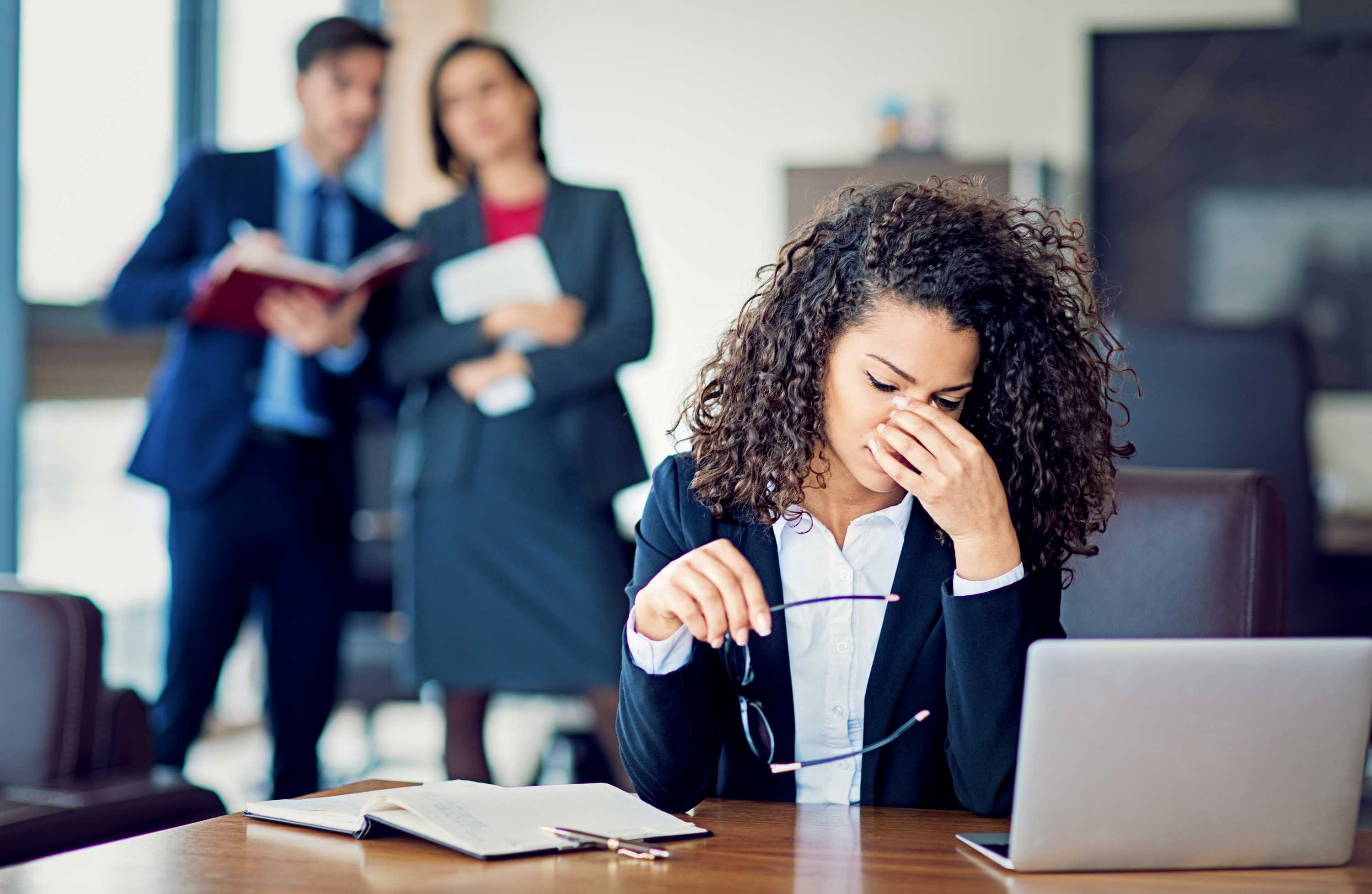 A woman sits at a desk looking worried while two figures stand behind her.