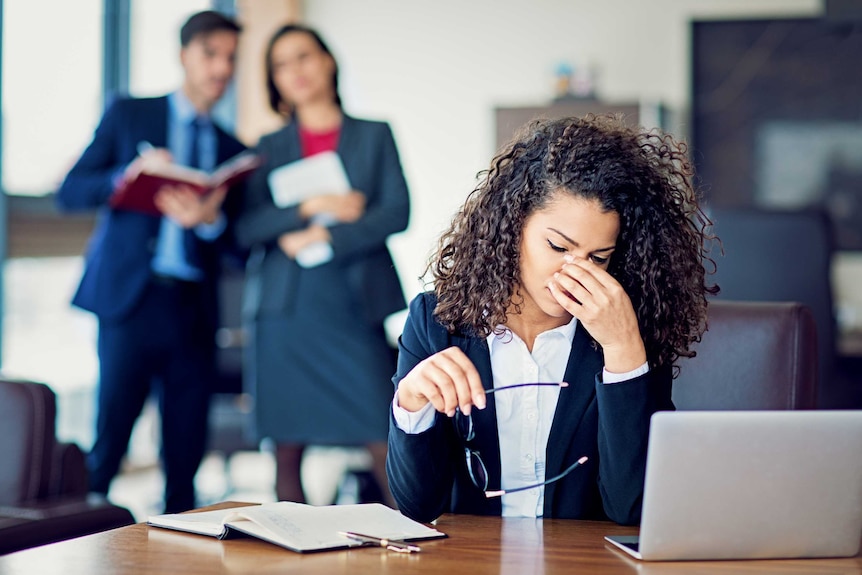 A woman sits at a desk looking worried while two figures stand behind her.