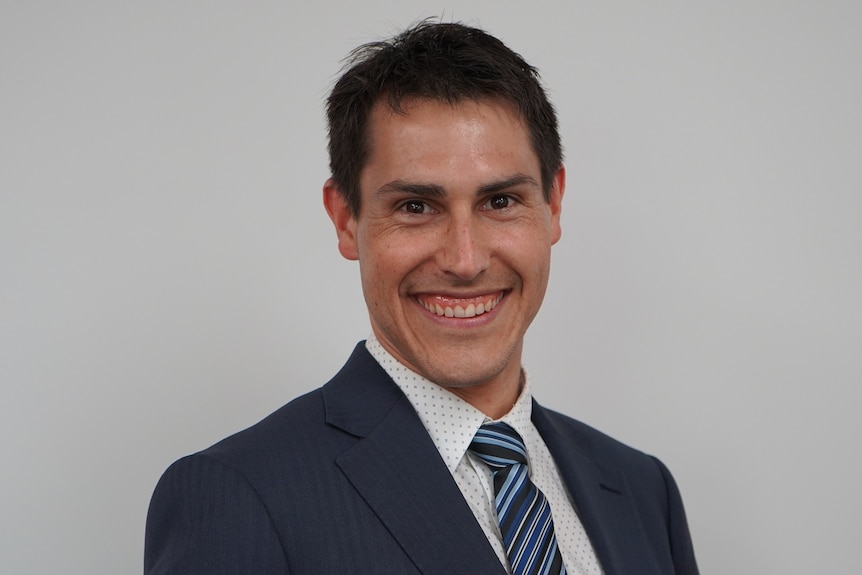 Luke Sartor wearing a dark suit, white shirt and striped blue tie, smiling at the camera.