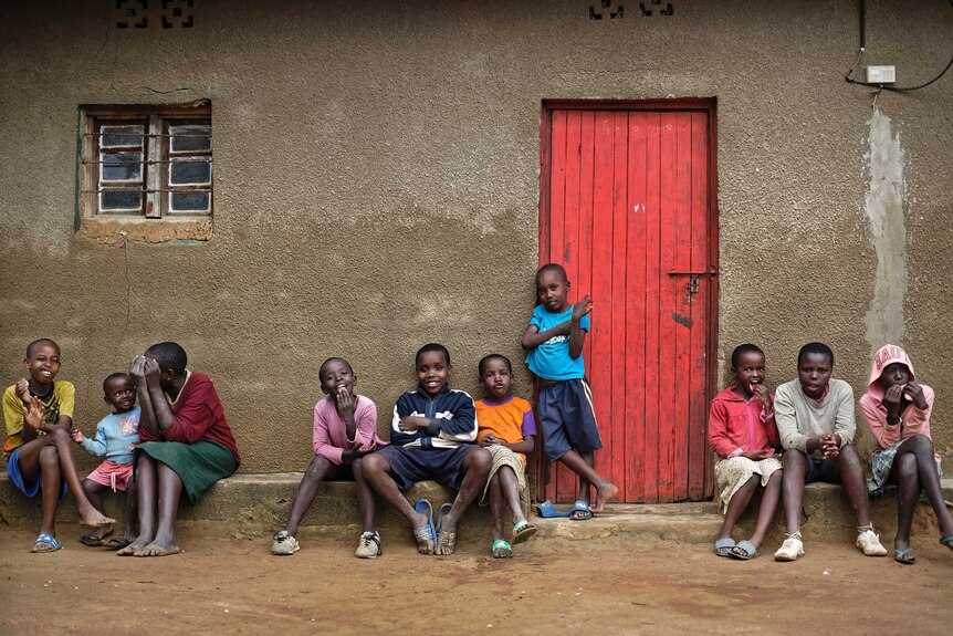 Ten children sit out the front of a house in front of a red door.