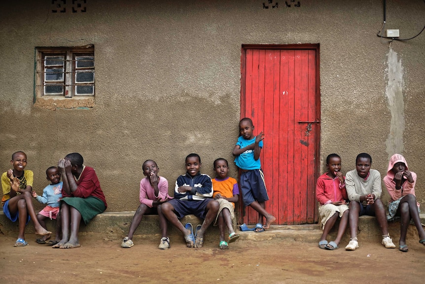 Ten children sit out the front of a house in front of a red door.