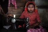 A woman with a hair covering and a red shirt works at a sewing machine while staring into the camera.