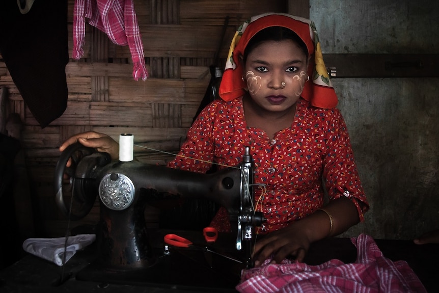 A woman with a hair covering and a red shirt works at a sewing machine while staring into the camera.