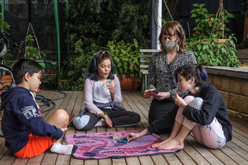 Emily Bieber on her back porch, playing uno with her 3 kids, sitting on wooden planks, green plants behind.