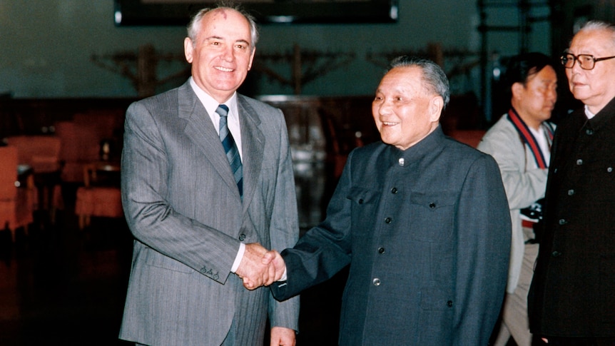 A man in a western suit grins as he shakes the hand of a man in a Mao suit