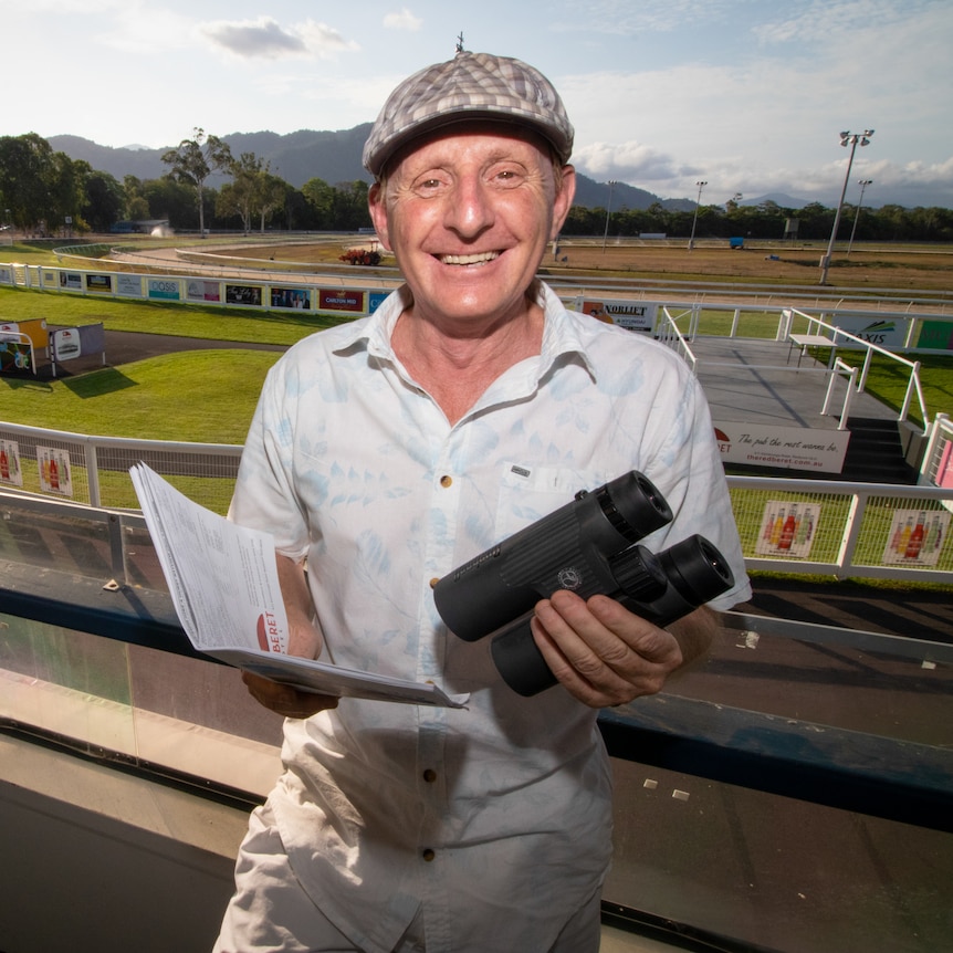 A man wearing a flat cap, holding a form guide and binoculars, in the stands at a racetrack.