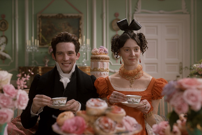 A man and woman in Regency England period costume each hold teacup and saucer and sit in ornately decorated period interior.