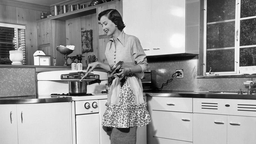 An black and white image of a woman from the 1950's in a kitchen wearing an apron and stirring a pot