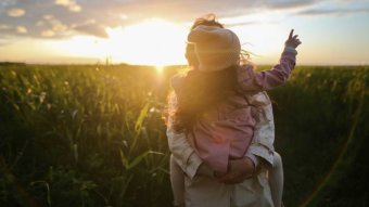 A child is sitting on her mother's back, pointing towards a beautiful sunset in a field of grass.