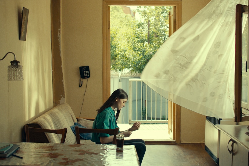 Young woman sits on couch in sunlit living room with gauzy window curtains blown towards her and open door behind her.