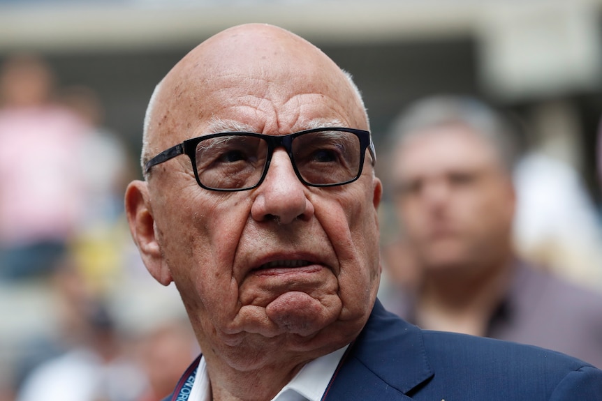 A  close up portrait of Rupert Murdoch, an elderly man with furrrowed features wearing glasses and a suit with open-necked shirt