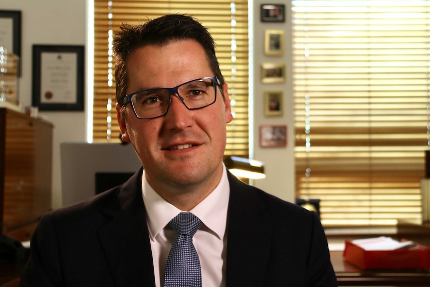 Zed Seselja, wearing glasses and a suit, sits at a desk in an office with sunlight pouring through the wooden blinds.