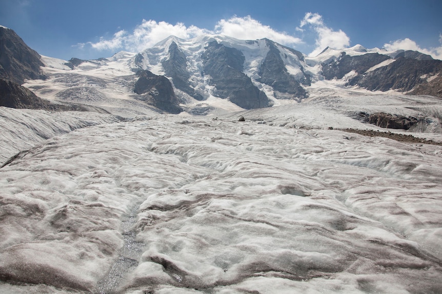An ice glacier with brown surface visable underneath and alps in the background. 