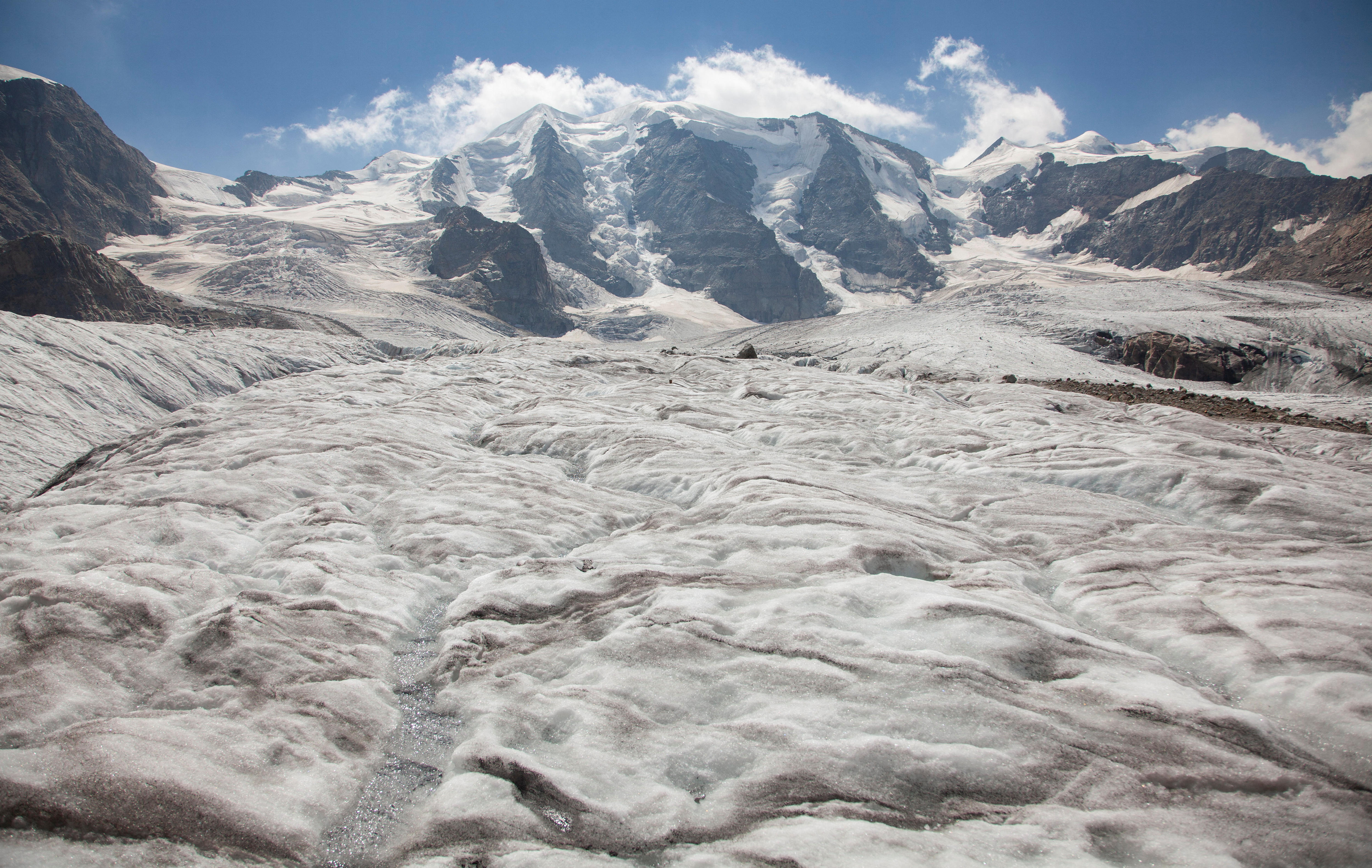 An ice glacier with brown surface visable underneath and alps in the background. 