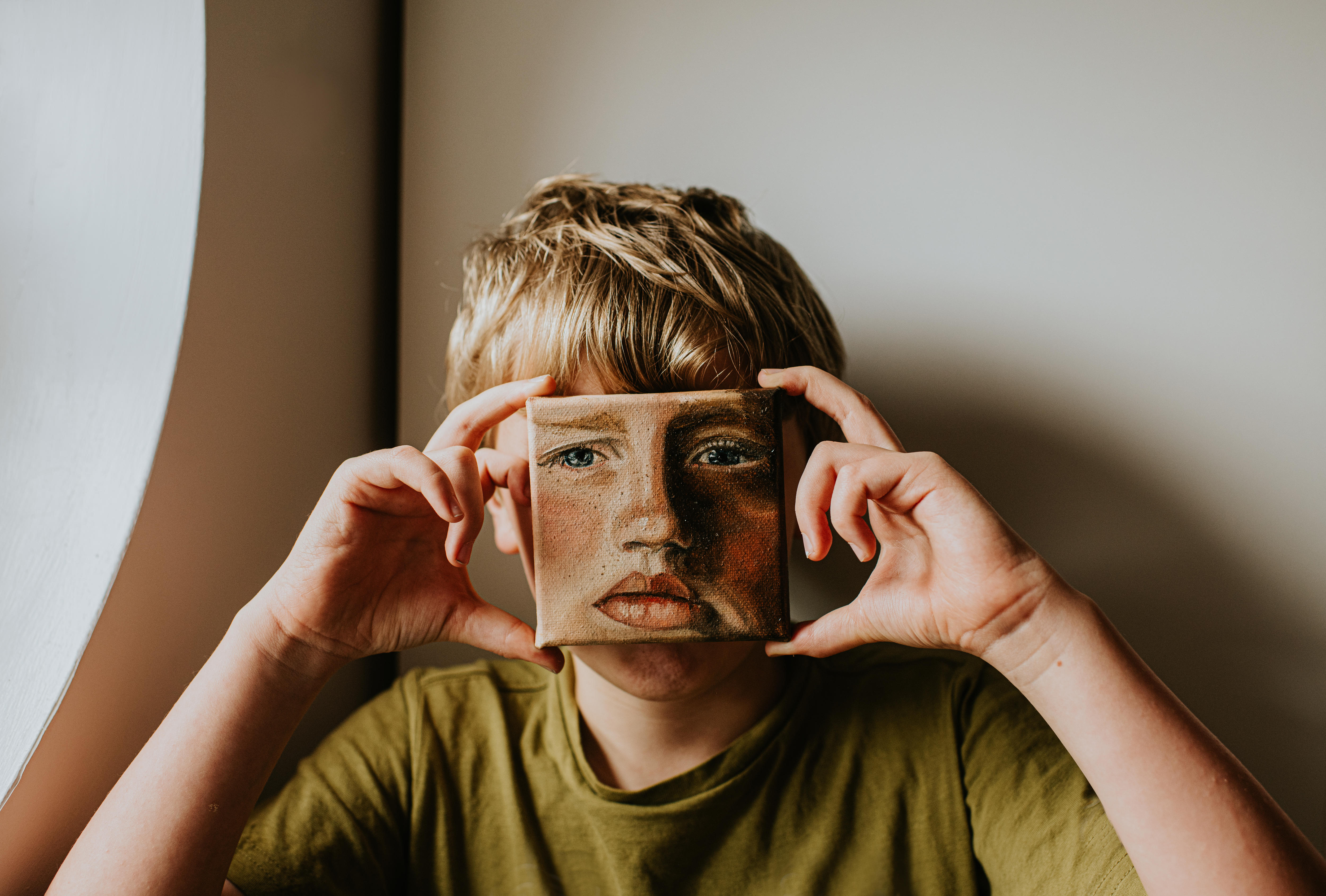 A young boy with blonde hair and a green t-short is holding up a small painted self portrait in front of his face