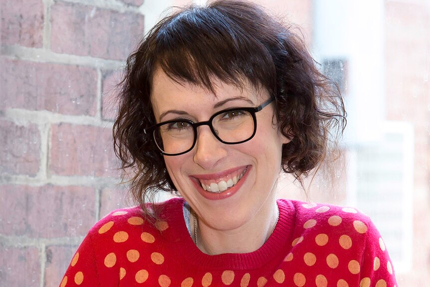 A smiling woman with short brown hair in a bright red jumper with black glasses.