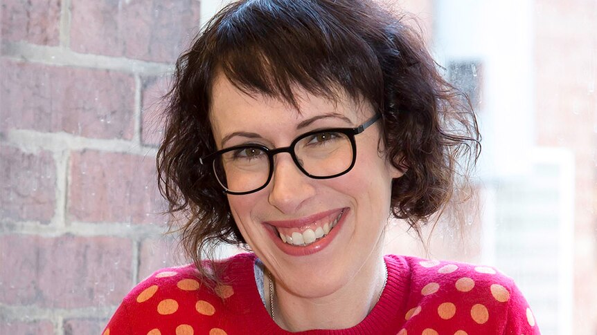 A smiling woman with short brown hair in a bright red jumper with black glasses.