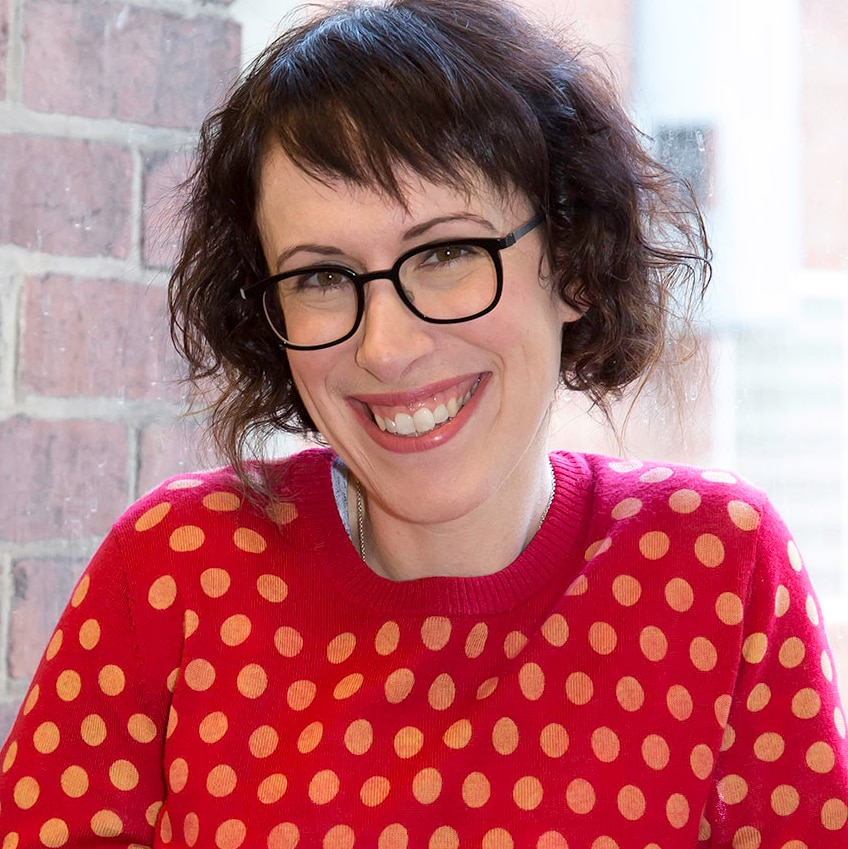 A smiling woman with short brown hair in a bright red jumper with black glasses.