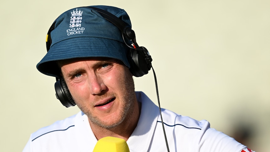 An England cricketer wearing a blue bucket hat and headphones over his ears speaks into a microphone. 