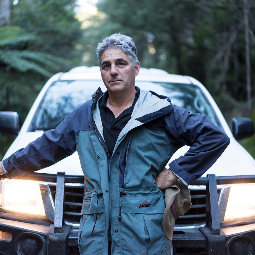 A man in a jumpsuit stands, leaning against a large car with headlights on, against the backdrop of a forest at dusk.