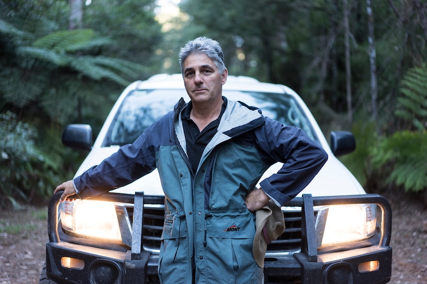 A grey-haired man in coveralls leans on a four-wheel drive in the bush.
