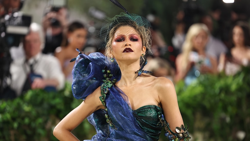 A mid shot of Zendaya in heavy makeup and a green and blue gown.