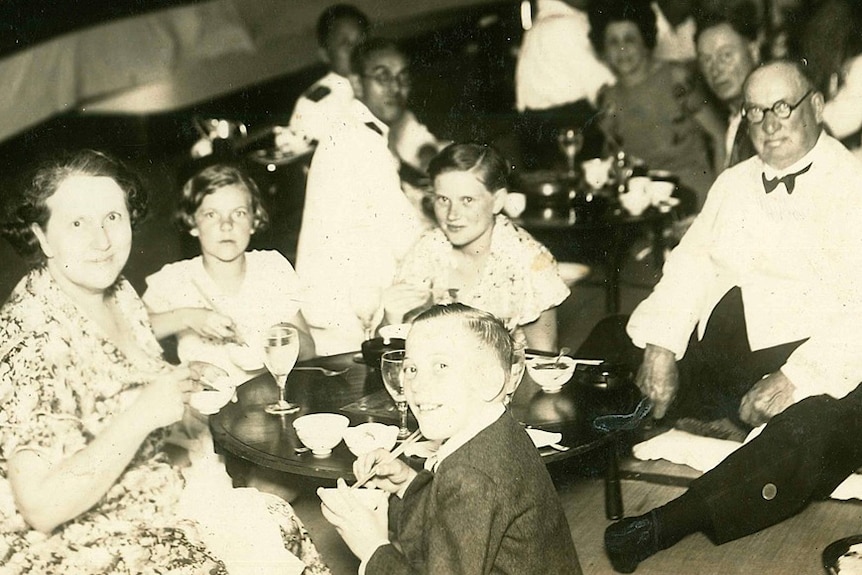 A family of two adults and three children sit at a table. looking towards the camera.