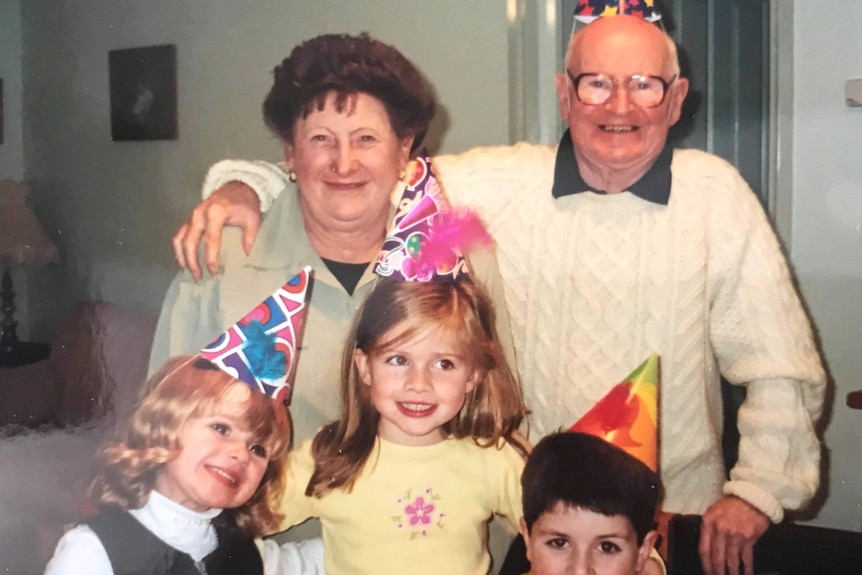 An elderly man and woman with their three young grandchildren all wearing party hats